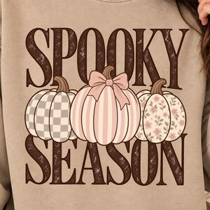 Könnte beinhalten: Ein beigefarbenes Sweatshirt mit Rundhalsausschnitt und dem Schriftzug SPOOKY SEASON in dunkelbraunen Buchstaben. Die Grafik zeigt drei Kürbisse: einen mit grau-weißem Karomuster, einen mit rosa Streifen und Schleife sowie einen mit rosa Blumenmuster.
