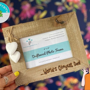 May include: A 6x4 inch driftwood photo frame with a rustic brown finish. The frame has a white heart hanging from it and the text "...World's Okayest Dad" is engraved on the bottom.