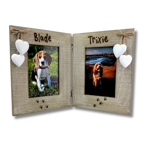 May include: A hinged, light-colored wooden picture frame with two openings, each displaying a dog photo. The frame is decorated with white heart ornaments and paw prints. The names "Blade" and "Trixie" are above each photo.