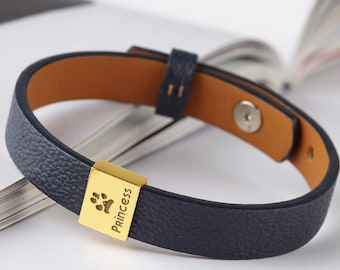 May include: A dark blue textured bracelet featuring a gold-toned rectangular metal charm engraved with a paw print and the word Princess. The bracelet has a tan interior lining, a small leather loop, and a visible silver-colored metal snap fastener.