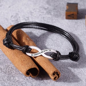 Può includere: Bracciale in pelle nera con un ciondolo infinito argentato inciso con coordinate. Il bracciale è annodato e ha un design semplice. Le coordinate sono 42.3593°N 85.8781°W.