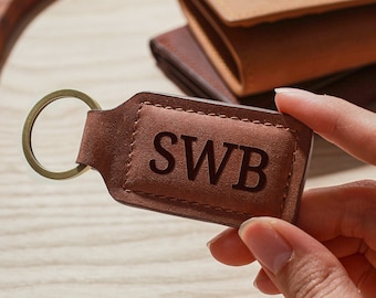 Op de afbeelding: Een bruine leren sleutelhanger met een gouden metalen ring en de initialen "SWB" gegraveerd in het leer.