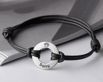 May include: A black cord bracelet with a silver circle charm engraved with a paw print and the name "Daisy".