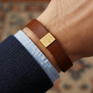 Op de afbeelding: Een bruine leren armband met een vierkant goudkleurig metalen middendeel. De metalen plaat is gegraveerd met een 'X'-vorm die de letters 'S', 'G', 'P' en 'M' in vier afzonderlijke kwadranten verdeelt. De band is glad en zit om de pols.