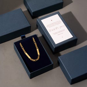 Puede incluir: Un collar de cadena dorada con placas con nombres, presentado en una caja de regalo azul marino con interior de terciopelo. La caja va acompa&ntilde;ada de una tarjeta de agradecimiento. El collar se muestra sobre una superficie gris.