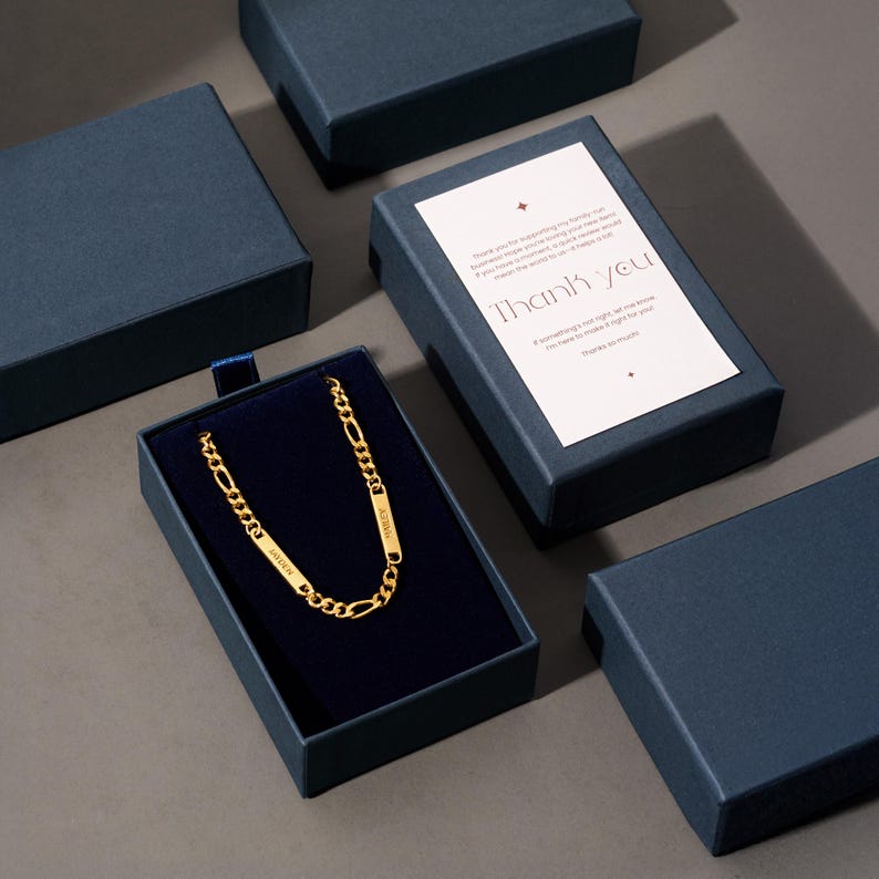 Puede incluir: Un collar de cadena dorada con placas con nombres, presentado en una caja de regalo azul marino con interior de terciopelo. La caja va acompa&ntilde;ada de una tarjeta de agradecimiento. El collar se muestra sobre una superficie gris.