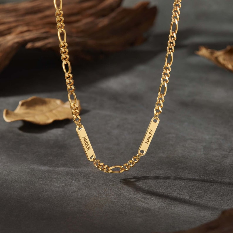 Puede incluir: Un collar de cadena Figaro dorado con dos placas rectangulares con nombres. Las placas tienen grabados los nombres "Jayden" y "Hailey". El collar se muestra sobre una superficie gris oscuro con un fondo borroso de madera y hojas.