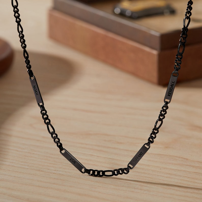 Puede incluir: Un collar de cadena figaro negro con cuatro placas rectangulares con nombres. Las placas est&aacute;n grabadas con los nombres "Noah", "Zoe", "Tiffany" y "Kendall". El collar se muestra sobre una superficie de madera clara.