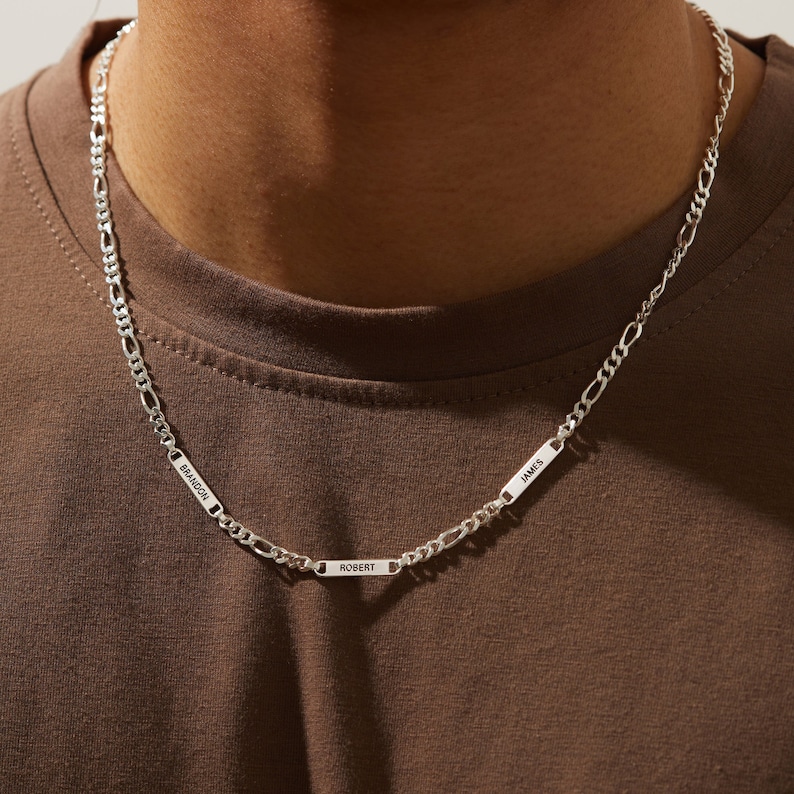 Puede incluir: Un collar de cadena Figaro plateado con tres placas rectangulares con nombres. Las placas con nombres est&aacute;n grabadas con los nombres "BRANDON", "ROBERT" y "JAMES". El collar se lleva alrededor del cuello.