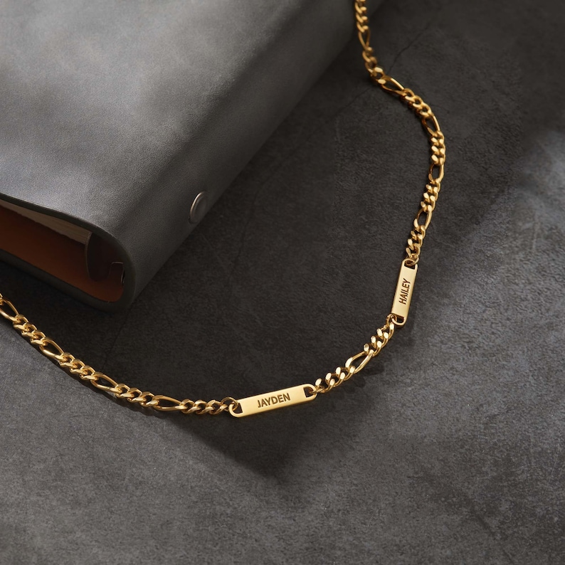 Puede incluir: Un collar de cadena Figaro de color dorado con dos placas rectangulares con nombres. Las placas con nombres est&aacute;n grabadas con los nombres "JAYDEN" y "HAILEY". El collar se muestra sobre una superficie gris oscuro junto a una cartera de cuero gris.