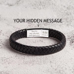 May include: A black braided leather bracelet featuring a silver-toned metal clasp. The clasp is engraved with the text "You're my always and forever" and "R&K 02.10.23". The text "YOUR HIDDEN MESSAGE" is printed above the bracelet.