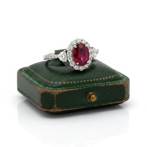May include: A silver-toned ring featuring a central oval-cut deep red ruby surrounded by a halo of small round white diamonds. The band is accented with additional diamonds and two pear-shaped stones on either side of the central setting.