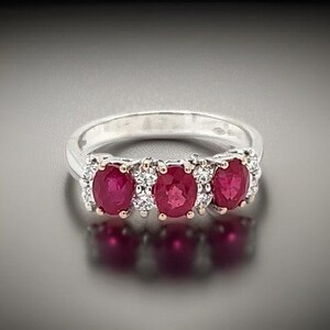 May include: A silver ring featuring three oval-cut rubies, each surrounded by small, sparkling diamonds. The rubies are a deep red color, and the ring has a classic, elegant design. The ring is displayed on a reflective surface.