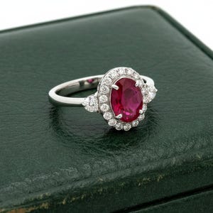May include: A silver-coloured ring featuring a central oval-cut deep red gemstone surrounded by a halo of small round white stones. The band is smooth and polished, with additional small white stones set into the shoulders of the ring setting.