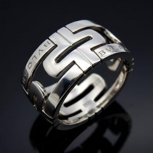 May include: A silver ring with a geometric design. The ring features the word "BVLGARI" engraved on the band. The ring has a polished finish and is displayed against a dark background.