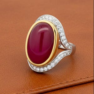 May include: A ring featuring a large, oval-shaped, deep magenta cabochon gemstone set in a yellow gold bezel. The band is crafted from silver-toned metal and is accented with a curved row of small, round, clear white sparkling stones.