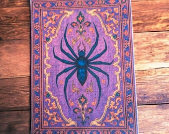 May include: A rectangular sticker featuring a stylized spider design. The spider is dark blue with a lighter blue center, set against a purple background. The border is ornate with orange and blue details, resembling a decorative rug.