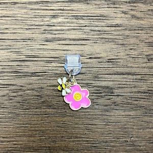 May include: A single earring with a pink flower and a small bee charm. The flower has a yellow center and gold trim. The bee is black and yellow with white wings. The earring post is clear plastic.