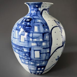 May include: A white ceramic vase with a blue and white geometric pattern. The vase has a wide base and a narrow neck.