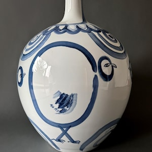 May include: A white ceramic vase with a blue and white hand-painted design of stylized birds. The vase has a round body and a narrow neck.