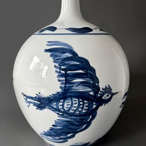 May include: A white ceramic vase with a blue and white bird design. The vase has a round body and a narrow neck.