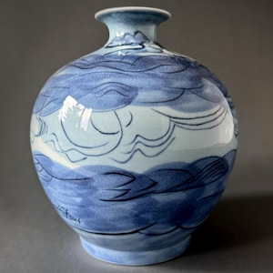May include: A blue and white ceramic vase with a rounded body and a narrow neck. The vase is decorated with a stylized design of clouds and waves. The vase is signed "Judith Freud".