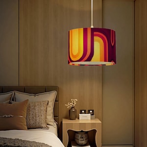 May include: A cylindrical pendant lamp with a retro design. The lampshade features a pattern of curved lines in shades of yellow, orange, and purple. The lamp hangs from a white cord in a bedroom setting.