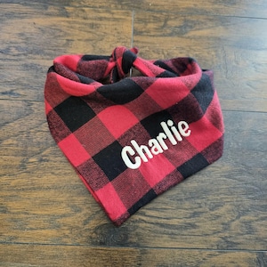May include: Red and black buffalo plaid dog bandana with the name "Charlie" in white script. The bandana is tied at the top and lies on a wooden surface. The fabric has a classic check pattern.
