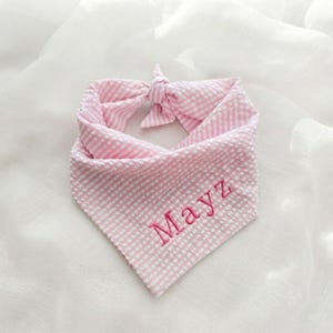 May include: A light pink and white gingham patterned pet bandana featuring a seersucker texture. The fabric is tied in a knot at the top, and the name Mayz is embroidered in dark pink thread across the lower front section of the triangular cloth.