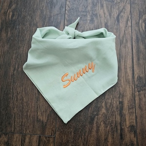 May include: A light green dog bandana with the word "Sunny" embroidered in orange. The bandana is knotted at the top and is made of a soft fabric. The bandana is laying on a wooden surface.