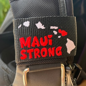 May include: A black scuba diving gear strap with a white patch that says "MAUI STRONG" in red letters and an outline of the Hawaiian islands in white.
