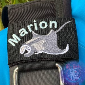 May include: Black fabric strap with the name "Marion" in light blue script and a gray stingray design. The strap is attached to a larger black object with a silver metal buckle. The background is a blurred mix of green and blue.