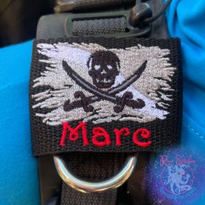 Puede incluir: Un parche bordado negro con un diseño de bandera pirata, que incluye una calavera y espadas cruzadas. La palabra "Mare" está bordada en rojo. El parche está sujeto a una correa negra con un anillo de metal plateado.
