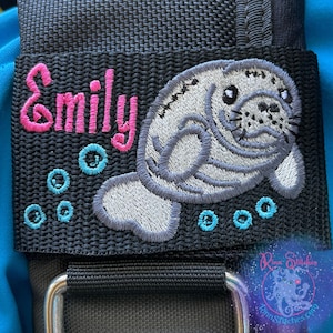 May include: Black fabric patch featuring the name "Emily" in pink embroidery, with a grey manatee design and blue embroidered bubbles. A silver metal buckle is visible at the bottom of the patch.