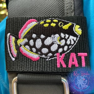 May include: Embroidered patch featuring a black and white fish with pink and yellow accents. The name "KAT" is embroidered in pink below the fish. The patch is attached to a black strap.