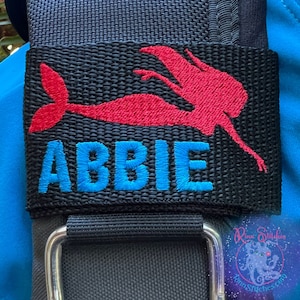 May include: Black strap with a red embroidered mermaid and the name "ABBIE" in blue embroidery.