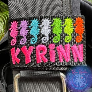 Puede incluir: Una correa de tela negra con un nombre bordado en rosa "KYRINN" y una fila de diseños de caballitos de mar coloridos. Los caballitos de mar son rosa, azul, blanco, verde y naranja.