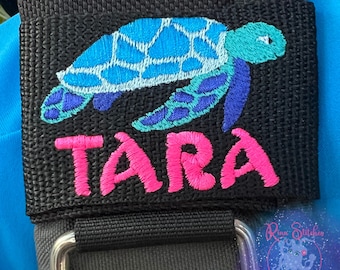 May include: A black fabric strap with a blue, green, and white embroidered sea turtle and the name "TARA" in pink embroidery.