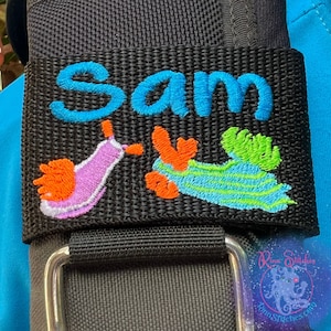 Puede incluir: Correa de tela negra con una hebilla plateada. La correa tiene un nombre bordado en azul "Sam" y dos babosas de mar bordadas de colores.