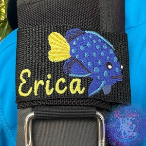 May include: A black fabric strap with a yellow embroidered name "Erica" and a blue and yellow embroidered fish. The strap has a silver buckle.