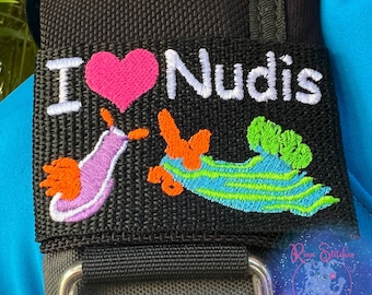 May include: Black fabric strap with a white embroidered heart and the text "I love Nudis". The strap also features two embroidered sea slugs, one purple and one green and orange.