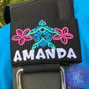 Puede incluir: Correa de tela negra con un diseño de tortuga bordada en rosa y azul y el nombre Amanda en letras blancas.
