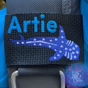 May include: Black adjustable strap with blue embroidered text "Artie" and a blue and white embroidered whale shark design.
