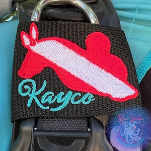 May include: Black strap with a red and white scuba diving flag and the name "Kayco" embroidered in turquoise.