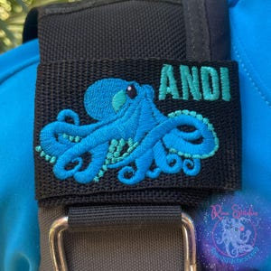 以下が含まれることがあります： 黒い生地のパッチに、ターコイズ色のタコ刺繍と「ANDI」という名前がターコイズ色で刺繍されています。タコの模様は詳細なステッチが施されています。パッチは、銀色の金属製の留め金が付いた黒いストラップに取り付けられています。