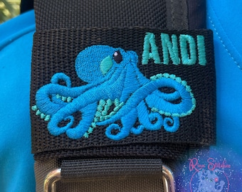 May include: Black fabric patch with a turquoise octopus embroidery and the name "ANDI" in turquoise. The octopus design features detailed stitching. The patch is attached to a black strap with a silver-colored metal clasp.