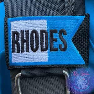 May include: Embroidered patch with the name "RHODES" in black block letters. The patch is divided diagonally, with a white left side and a blue right side. The patch is sewn onto a black strap.