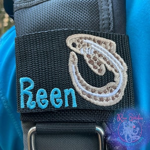 May include: Black strap with the name "Reen" embroidered in turquoise. A beige and brown embroidered fish design is on the right side. The strap is attached to a gray harness with a silver metal buckle.