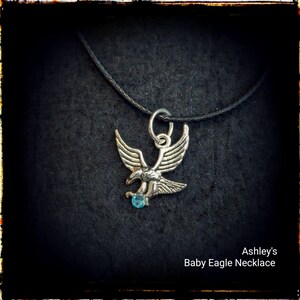 May include: A silver baby eagle necklace with a blue gemstone on a black cord. Ashley's Baby Eagle Necklace.
