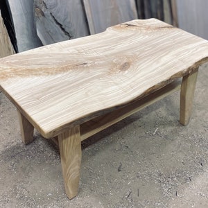 Könnte beinhalten: Ein Couchtisch aus hellem Holz mit einer Live-Edge-Oberfläche und einem schlichten, modernen Design. Der Tisch hat eine natürliche Holzoptik und wird von vier Beinen getragen.